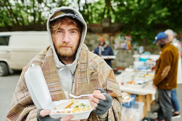 A man eating food outside, near a group of individuals serving food to those experiencing hunger