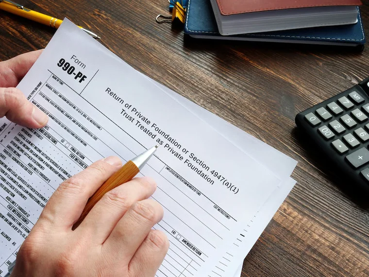An individual sits at a desk, pen in one hand and the IRS Form 990 in the other.