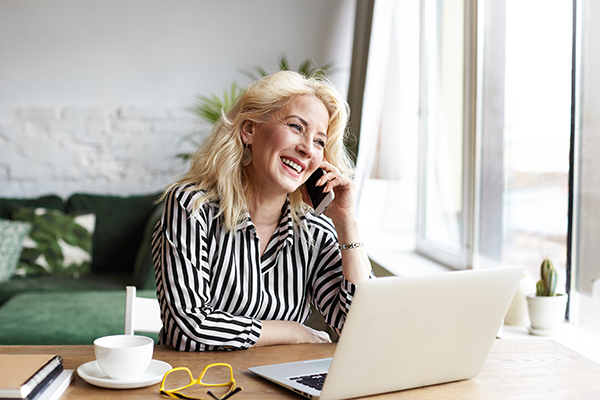 A woman smiles in front of her laptop while on the phone.