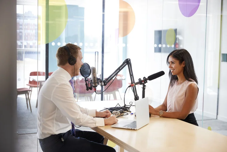 Two media professionals sit as a desk with microphones having a discussion