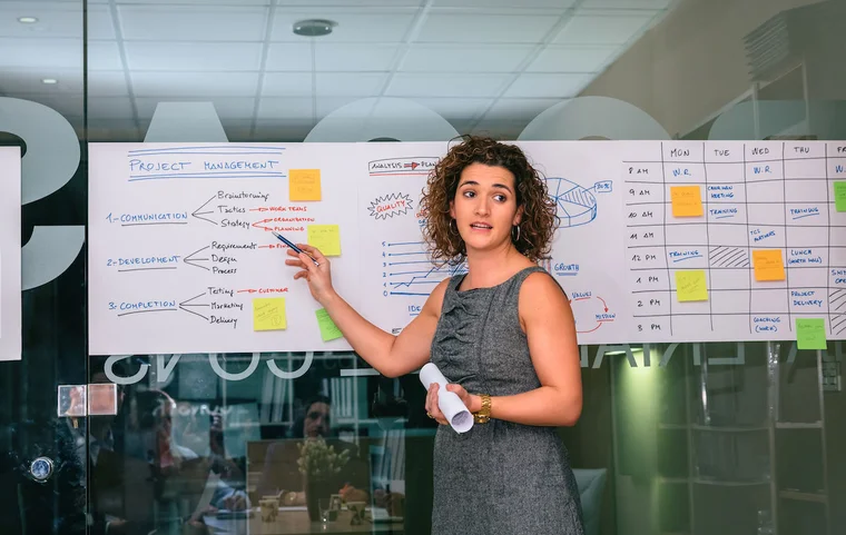A woman stands in front of a white board with sticky notes and project plans