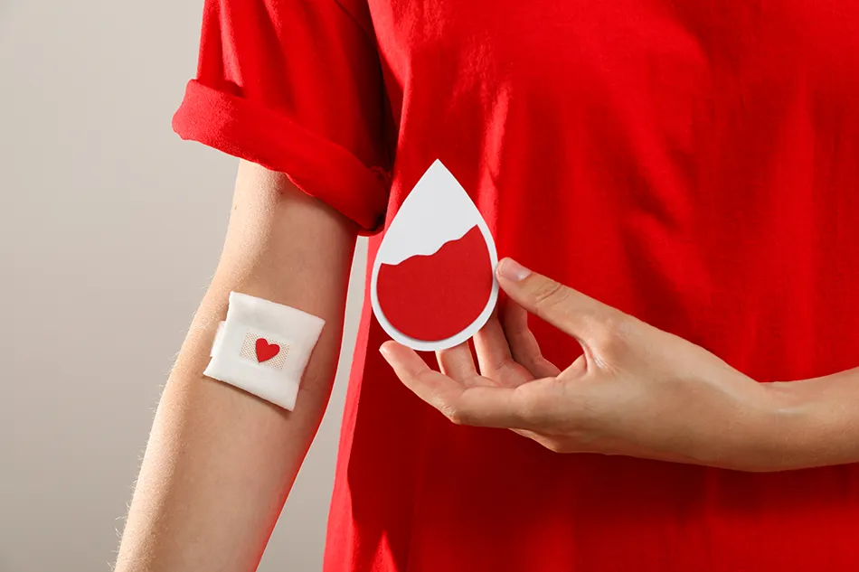 A image of a bandaid on the inside of an individuals arm with a heart after giving blood, he holds a paper representation of a blood drop