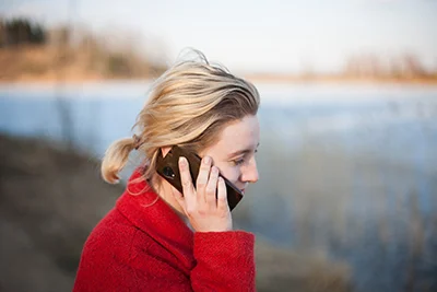 A woman in a red jacket holds a phone to her year outside near a lake
